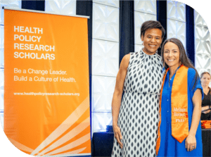 Keshia and Melissa Horner at the 2024 HPRS Graduation during Summer Institute. They are standing to the right of a HPRS banner smiling. Melissa is wearing an orange graduation stole.