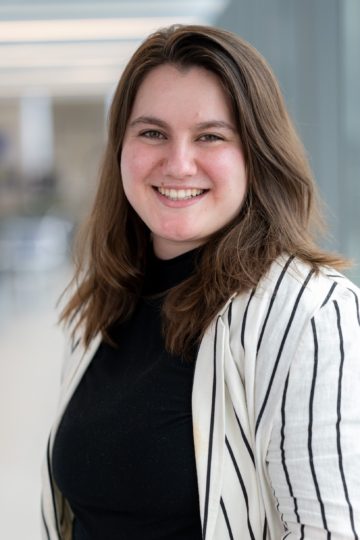 The image shows a person smiling warmly, with shoulder-length brown hair, wearing a black top and a white blazer with thin black stripes. They are positioned against a blurred indoor background with soft lighting, giving the photo a professional and approachable feel. The person has a friendly expression and is looking directly at the camera.