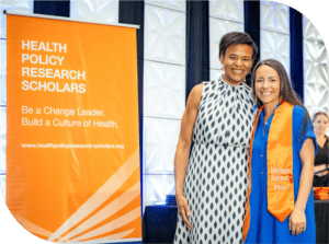 Keshia and Melissa Horner at the 2024 HPRS Graduation during Summer Institute. They are standing to the right of a HPRS banner smiling. Melissa is wearing an orange graduation stole.